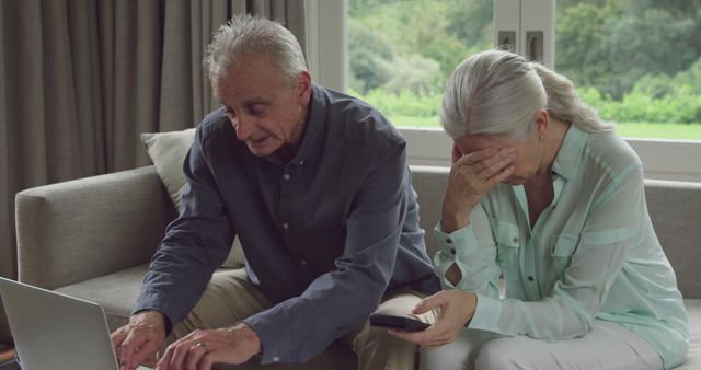 Senior Caucasian couple looks at a laptop in a home setting. They appear concerned or troubled, dealing with bad news or a difficult situation.