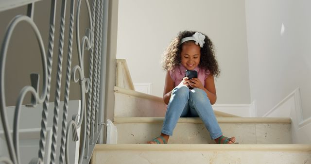 A young girl is sitting on stairs at home, looking excited while using a smartphone. The child, with her curly hair adorned with a white bow, is dressed casually in a pink top and jeans, reflecting a relaxed and enjoyable moment. This can be used for depicting modern childhood, technology use, family life, or promoting home safety and comfort.