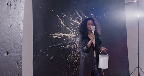 African American Woman Holding Paintbrush and Bucket in Studio with White Paint Splash