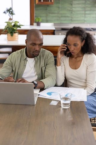 Diverse couple reviewing finances and charts at home using laptop and phone