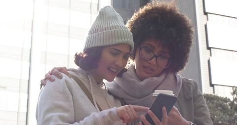 Young diverse women sharing smartphone and coffee on sunlit city plaza, pointing and smiling