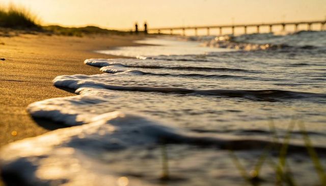 Golden hour surf lapping wet sand with wooden pier and walking silhouettes at shoreline