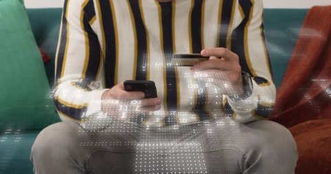 Middle-aged man holding smartphone and tapping credit card on couch with digital overlay