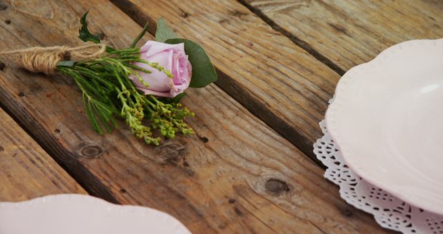 A rustic table setting features a delicate pink rose bouquet next to elegant white plates with a lace detail, with copy space. The wooden texture of the table adds a warm, natural feel to the romantic or celebratory occasion.