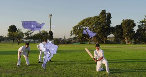 Kneeling batsman preparing to hit with teammates fielding on sunlit park grass at golden hour