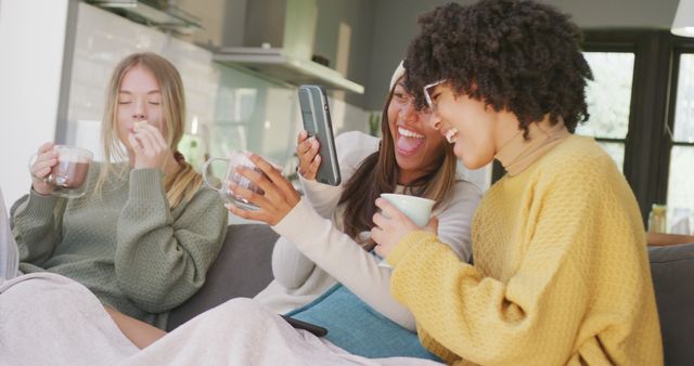 Happy diverse teenager girls sitting on sofa, drinking cocoa and using smartphone. Spending quality time, lifestyle, friendship and adolescence concept.