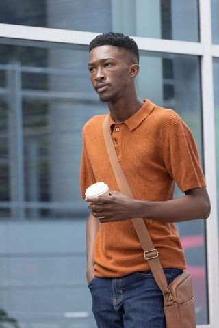 Young african man standing with coffee cup and crossbody bag by glass facade