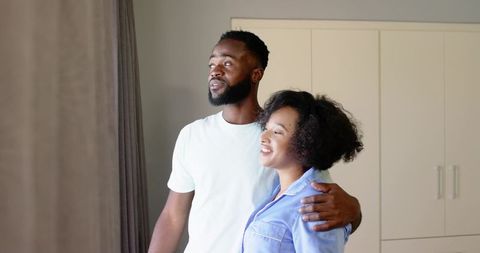 Mid-adult couple embracing and looking out sunlit window, cozy bedroom moment