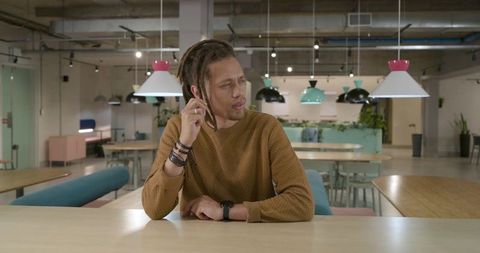 Thoughtful man sitting at communal table in coworking space touching ear, modern industrial office