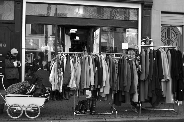 Vintage clothing store front with racks of clothes and two shoppers. Black and white image captures outdoor retail experience with retro aesthetics. Perfect for use in articles about fashion trends, retail businesses, second-hand shopping, and urban street activities.
