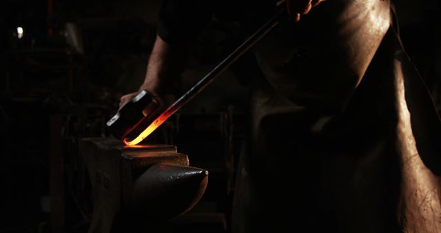 Blacksmith at work hammering glowing hot metal on an anvil in a dimly lit workshop. Ideal for use in articles on artisanal crafts, manual labor, traditional skills, and metalworking tutorials.