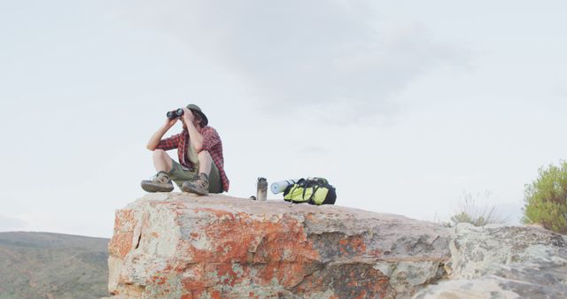 A man is sitting on a rocky cliff in an outdoor setting, using binoculars to look into the distance. He is accompanied by a backpack and a thermos, suggesting a hiking or adventure trip. This image is perfect for use in content related to outdoor activities, adventure travel, hiking tips, nature exploration, and leisure activities.