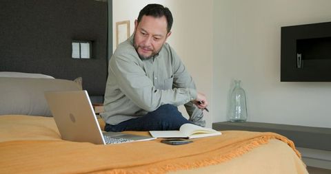 Asian Man Focusing on Laptop in Cozy Bedroom Workspace