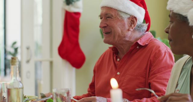 Diverse senior man and woman in santa hats listening and laughing at christmas dinner table at home. retirement lifestyle, christmas festivities, celebrating at home with friends.
