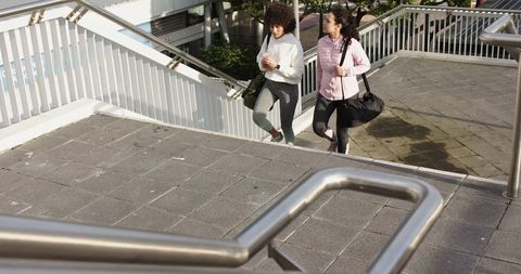 Two women walking up overpass stairs carrying gym bag and checking smartphone in city