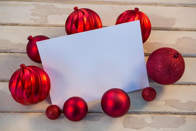Blank card surrounded by red Christmas baubles on wooden surface. Ideal for holiday greeting cards, festive invitations, Christmas promotions, and seasonal advertisements.
