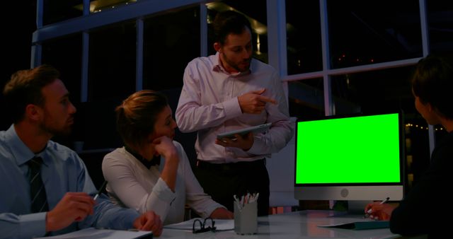 This image shows a professional business team analyzing data on a computer with a green screen in an office during nighttime. Perfect for illustrating themes of teamwork, technology, and business strategy. Suitable for articles, blogs, website headers, and presentations related to corporate work environments, digital marketing, and collaborative projects.