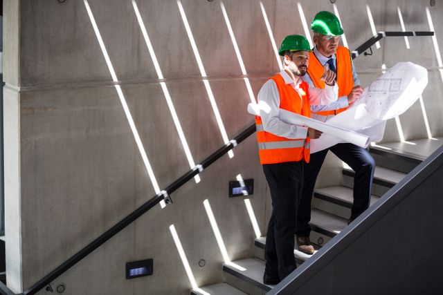 Architects in safety vests and hard hats are discussing a blueprint while standing on a staircase in a modern office building. This image can be used to illustrate themes of construction, teamwork, project planning, and architectural design. It is suitable for use in articles, presentations, and promotional materials related to engineering, architecture, and construction projects.