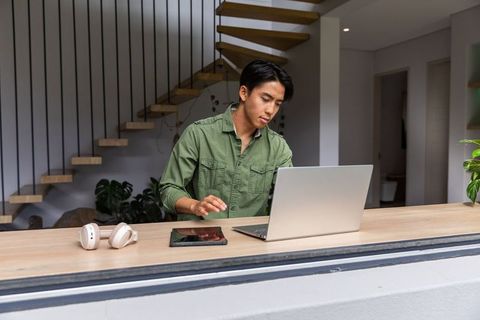 Asian man working remotely using laptop and tablet in modern open-plan home office