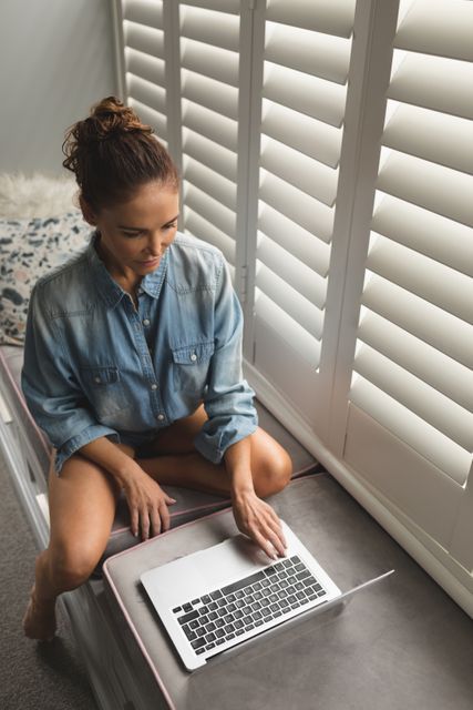 High angle view of a woman sitting on a window seat using a laptop. She is wearing a denim shirt and appears relaxed and focused, surrounded by natural light filtering through the shutters. This image can be used for themes related to remote work, comfortable home living, lifestyle, or technology.