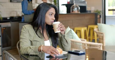 Mid-adult Indian woman sipping coffee and holding pastry at modern cafe table with phone