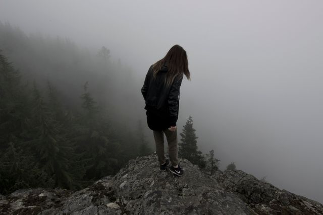 Person standing on a rocky cliff surrounded by a misty forest. Perfect for use in campaigns related to adventure, outdoor activities, and exploration. It can evoke feelings of solitude, serenity, and connection with nature.