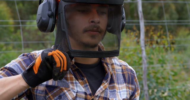 A young Latino man wears safety gear, including a face shield and ear protection, while adjusting his equipment, with copy space. His attire suggests he is preparing for or is engaged in work that requires personal protective equipment.