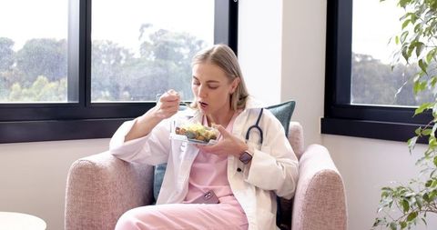 Female clinician enjoying healthy lunch in clinic lounge wearing coat and stethoscope