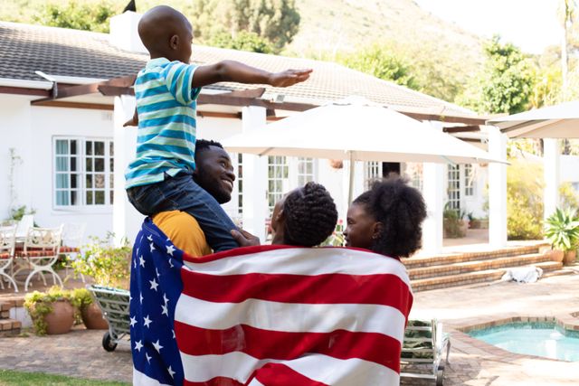 This image shows a joyful African American family in their backyard, holding an American flag. The son is on his father's shoulders with his arms spread wide, symbolizing freedom and happiness. This image is perfect for themes related to family bonding, patriotism, celebrations, and domestic life. It can be used in advertisements, social media posts, and articles about family values, national pride, and summer activities.