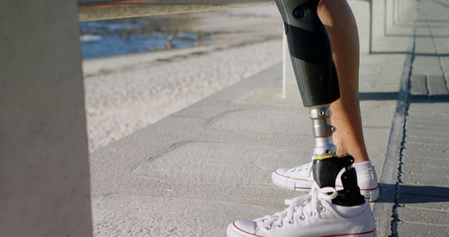 Shows person wearing sneakers and using prosthetic leg standing at beach, perfect for illustrating themes of overcoming obstacles, mobility, and freedom. Useful for websites, articles, and campaigns focusing on health, ability, and summer activities by the seaside.
