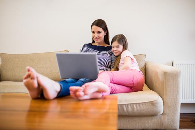 Mother and daughter sitting on a sofa using a laptop together in a cozy living room. Perfect for themes related to family bonding, technology use at home, parenting, and leisure activities. Ideal for articles, blogs, and advertisements focusing on family life, home comfort, and digital learning.