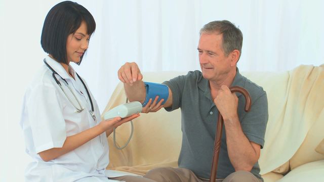 Nurse is checking the blood pressure of an elderly male patient who is sitting on a couch holding a walking stick. This scene is perfect for depicting home healthcare services, medical insurance ads, or elder support articles. It highlights quality caregiving and the importance of health assessments for seniors.