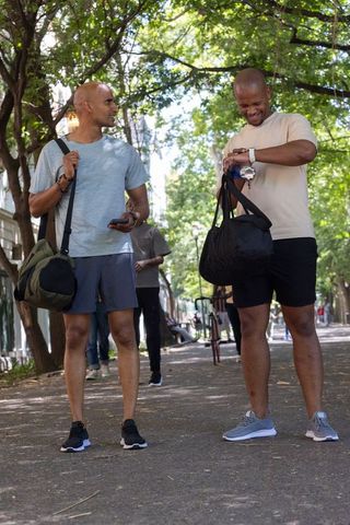 African american friends walking in tree-lined park carrying gym duffels checking watch