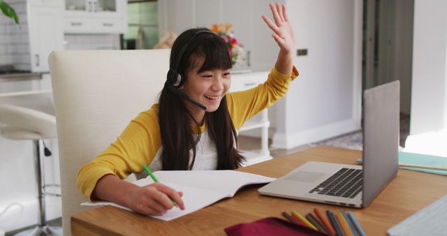 Asian girl wearing a yellow shirt smiling while participating in an online class at home. She is using a headset and looking at her laptop, taking notes in a notebook with colored pencils on the table. Suitable for educational materials, remote learning content, and articles about online education.