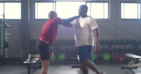 African men sparring with knee strike in industrial gym near dumbbell rack, backlit