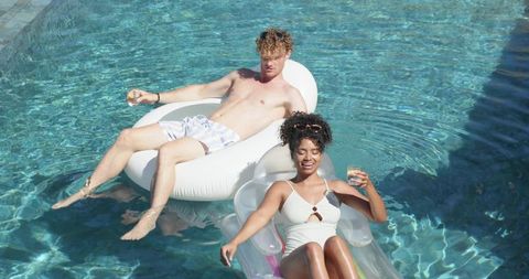 Couple relaxing on inflatable floats in sunlit pool sipping cocktails and enjoying summer