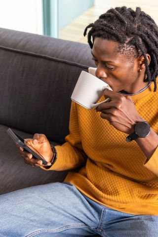 African american man sipping coffee and checking smartphone on sofa in mustard sweater