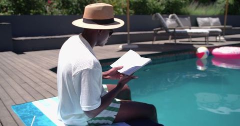 African man reading by pool in straw hat, relaxing on towel beside modern backyard pool