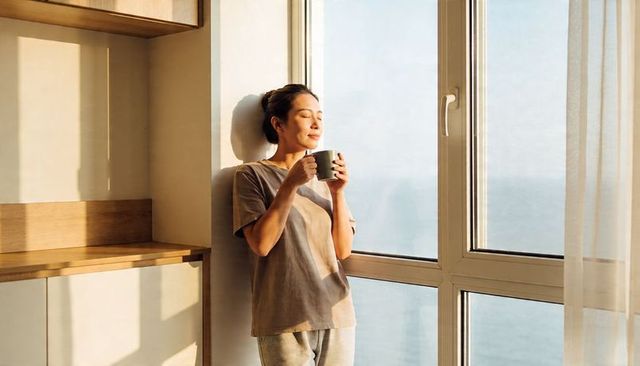 Asian woman enjoying morning coffee by sunlit window with sea view in minimalist kitchen