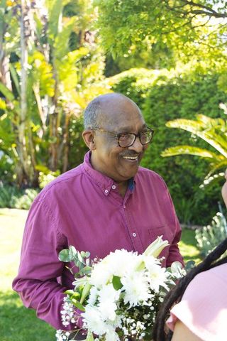 Smiling african american senior man holding white flower bouquet in sunny backyard garden