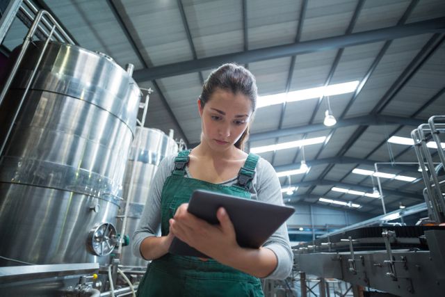 Female worker in uniform using tablet in a modern beverage production facility. Stainless steel tanks and machinery are visible in the background. Useful for illustrating industrial work environments, technology in manufacturing, quality control processes, and modern factory operations.