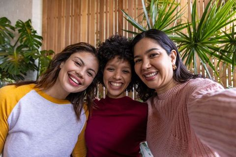 Three friends smiling and taking selfie in cozy cafe with plants, wood tones, casual lifestyle
