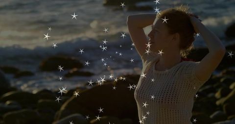 Woman gathering hair on rocky shoreline at sunset with star sparkles and golden-hour glow