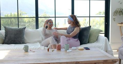 Female friends relaxing with eye patches on sofa, home spa skincare routine in sunlit room