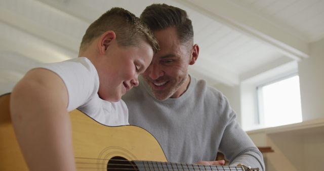 Father teaching his son to play the guitar indoors, creating an intimate and positive bonding moment. They are smiling and supportive, symbolizing happy family time and parenting. Suitable for content related to family relationships, music education, and positive parenting.