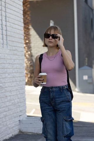 Young woman walking with coffee and sunglasses on urban sidewalk in denim cargo pants