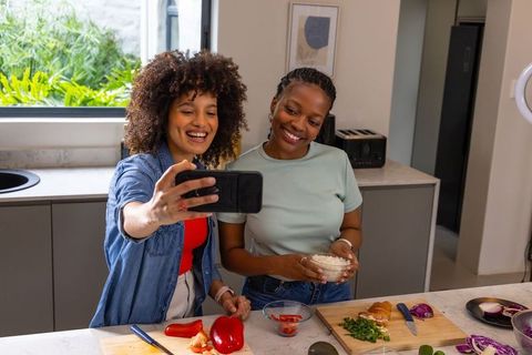 Diverse friends taking selfie while preparing healthy meal on sunlit kitchen island