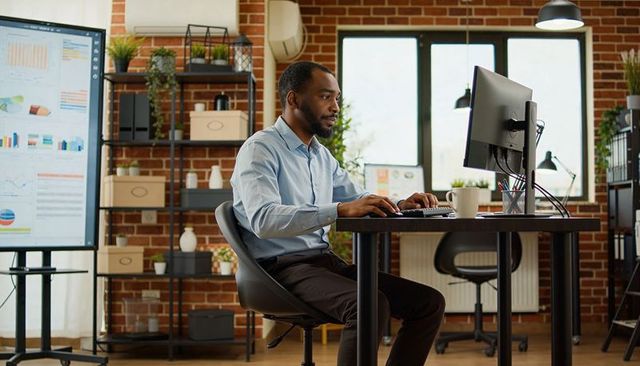 African american professional typing at modern brick office desk analyzing data on monitor