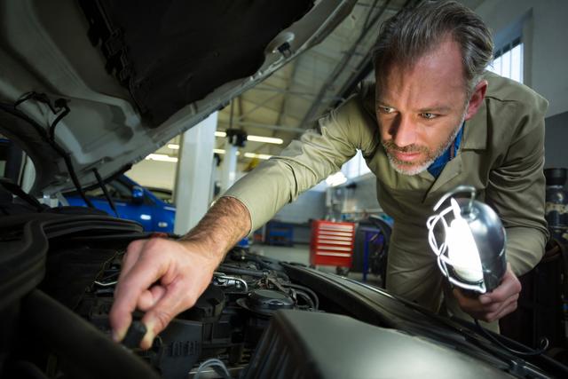 Mechanic inspecting car engine with a lamp in a repair garage. Useful for illustrating automotive repair services, professional mechanics at work, and vehicle maintenance. Ideal for websites, advertisements, and articles related to car repair, auto workshops, and mechanical services.