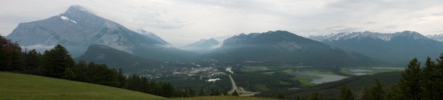 Panoramic view of a majestic mountain landscape with a town nestled in the valley. Dark stormy clouds loom over the textured peaks, while a highway leads through the landscape. Ideal for use in travel blogs, tourism brochures, outdoor adventure magazines, and nature-themed content.